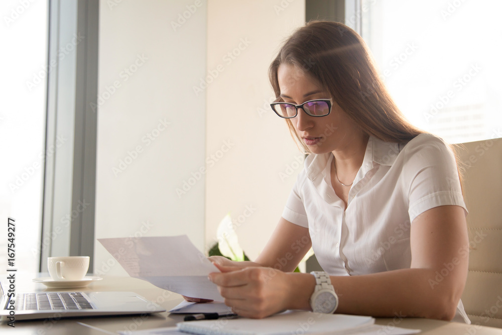 Serious worried businesswoman attentively reading letter with bad news sitting at work desk, anxious woman holding document, looking at declined rejected loan application or dismissal notification