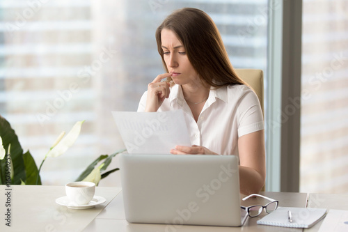 Concentrated focused businesswoman holding document, attentively reading financial report, analyzing work results, thinking over project management statistics, ways to improve business strategy