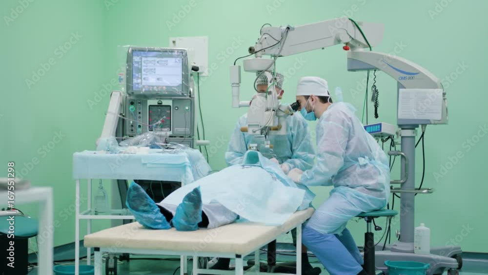 Team of ophtalmology surgeons and assistants with ophtalomological surgery tools during surgical treatment. Two male surgeons operate the machinery. Indoor. Wide angle. Stabilised Team of ophtalmology surgeons and assistants with ophtalomological surgery tools during surgical treatment. Two male surgeons operate the machinery. Indoor. Wide angle. Stabilised