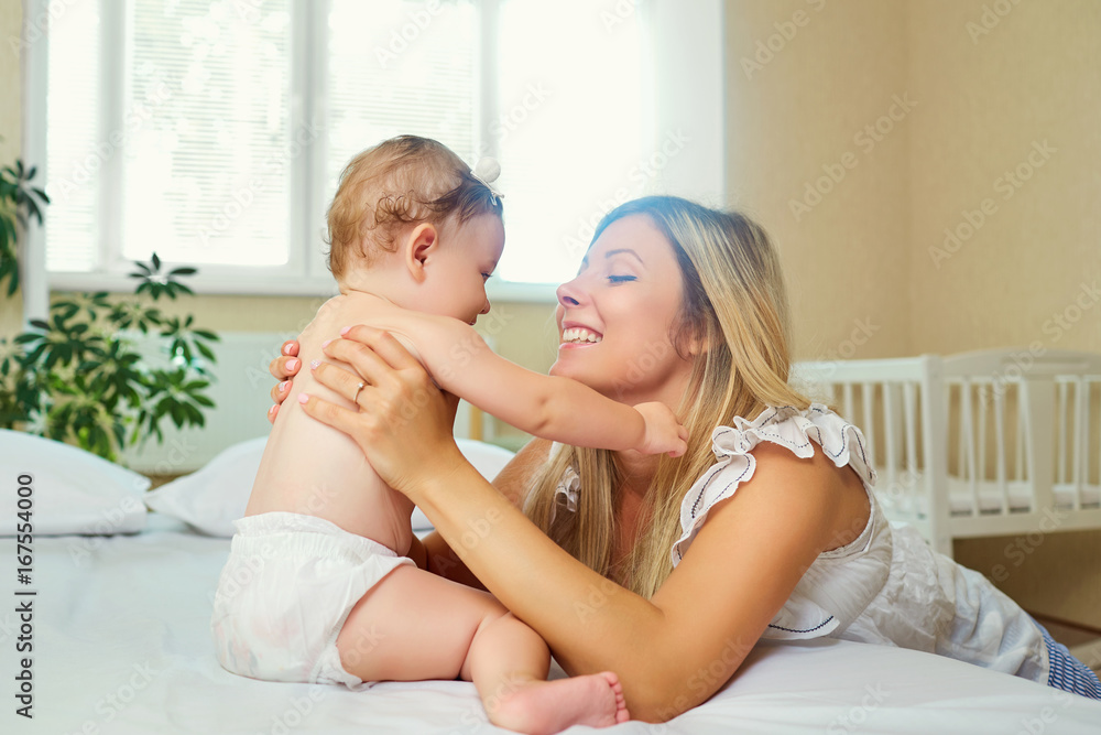 Mother and baby in a diaper play hugging on a bed indoors. Mother and baby in a diaper play hugging on a bed indoors.