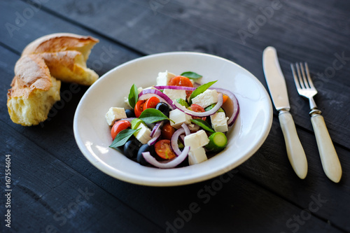 fresh and healthy vegetable mediterranean salad with feta cheese, cucumber, tomato, olives and red onion bulbs. ingredients and fresh baked homemade greek country bread on background, close-up