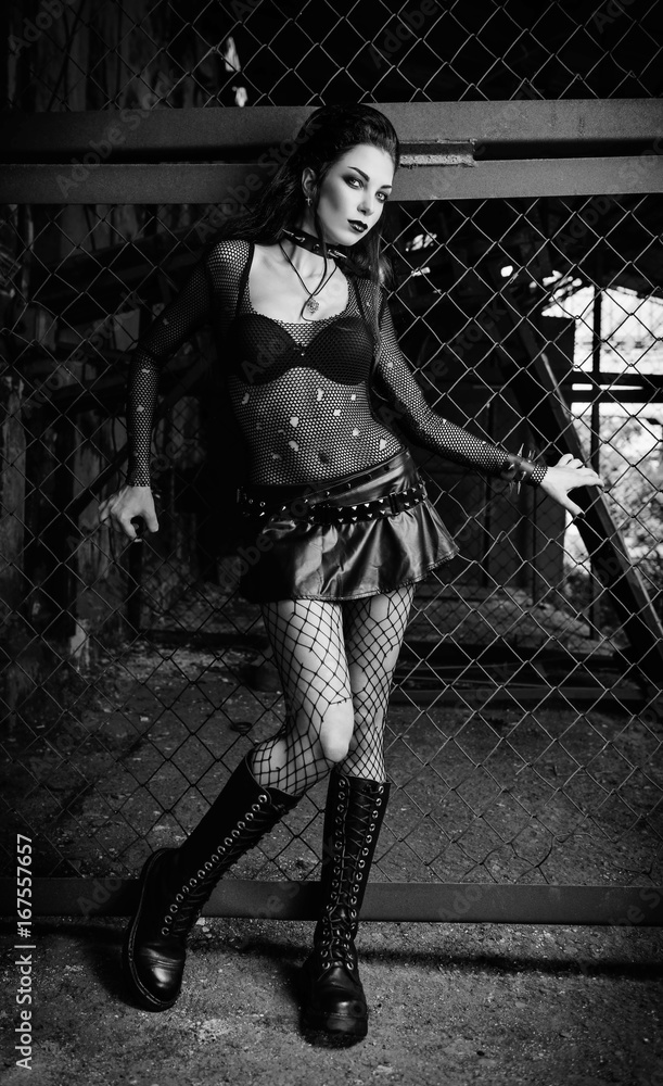 Foto Stock Portrait of sexy goth (deathrock) girl dressed in holey blouse,  skirt, bra and boots standing in industrial place. Black and white | Adobe  Stock