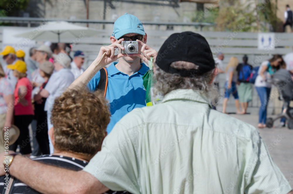A young man taking a snapshot of his elderly parents on a tourist site. Indistinctive tourists on the background.