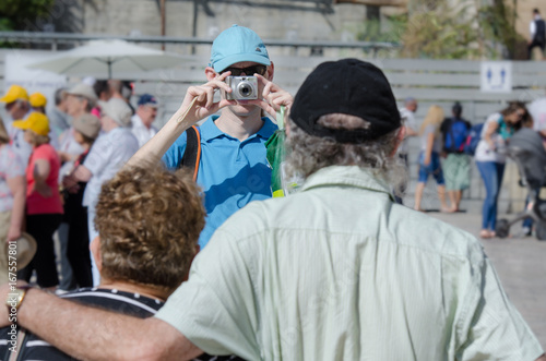 A young man taking a snapshot of his elderly parents on a tourist site. Indistinctive tourists on the background.