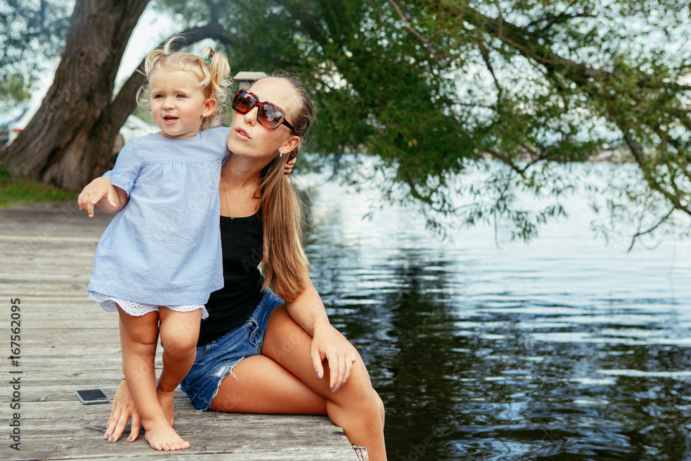 Fototapeta premium Group portrait of happy white Caucasian mother and daughter child having fun outside. Mom and child girl playing, hugging in park on pier by water lake. Candid authentic real lifestyle.