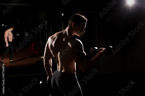 Handsome power athletic guy bodybuilder doing exercises with dumbbell. Fitness muscular body on dark background.
