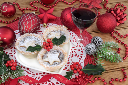 Christmas mince pie cakes on a heart shaped plate with dessert wine, holly, fir and red bauble decorations on a place mat on oak table background.