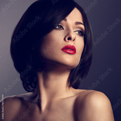 Beautiful makeup woman with perfect face, short black hair style and red lipstick on dark shadow background looking serious. Closeup toned portrait