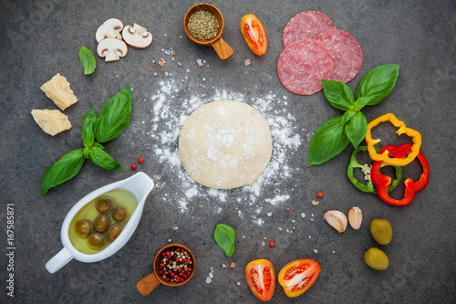 Raw dough for homemade pizza with ingredients and herbs  flat lay on dark stone background.