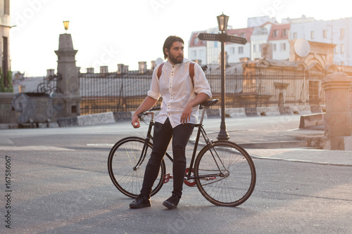 Young man posingwith the bicycle in city, hipster with leather backpack and fixie bike , sunrise background 