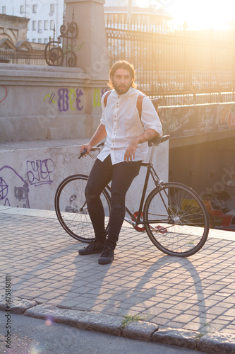 Young man posingwith the bicycle in city, hipster with leather backpack and fixie bike , sunrise background   photo