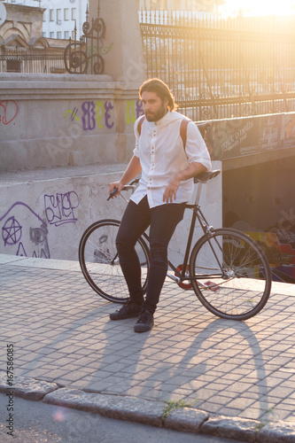 Young man posingwith the bicycle in city, hipster with leather backpack and fixie bike , sunrise background   photo