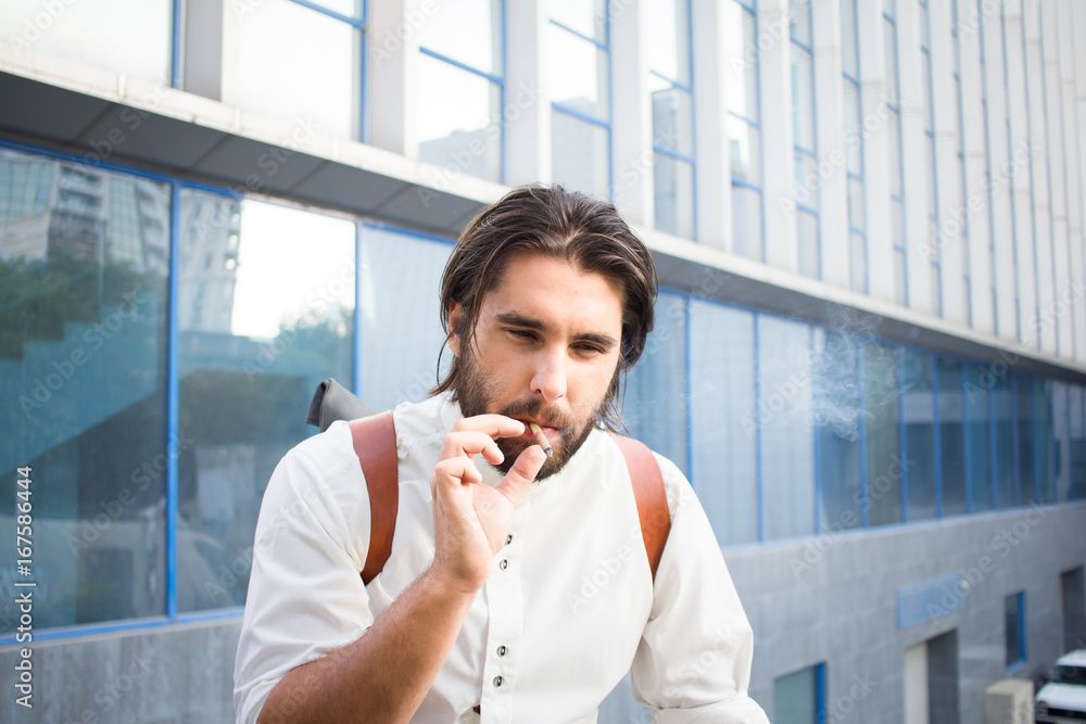 handsome guy with beard smocking cigarette on urban background with blue and gray glass  