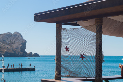 View at the part of balcony decorated with a fishnet with starfishes. Sea with mountain and pier with peoples on the background. Sudak, Crimea