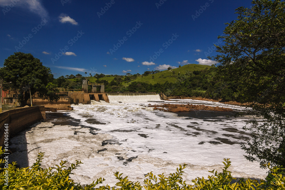 A água poluída passando pela barragem, tendo como possível causa os esgotos sem tratamento liberados no rio em contraste com a bela paisagem