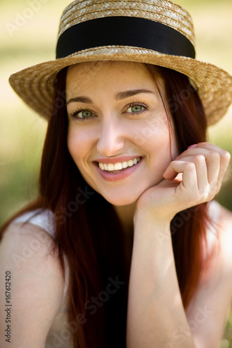 Portrait of young foxy lady in garden. Close up view of beautiful woman in straw hat, holding her hand close to chin.