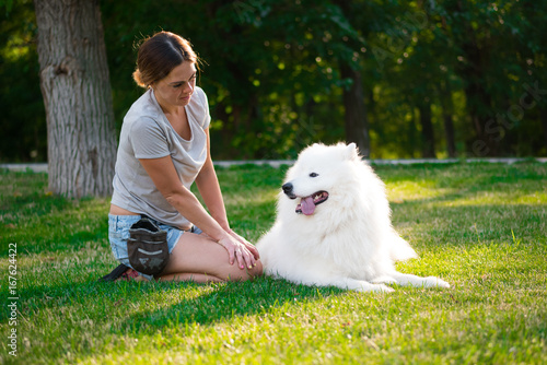 An adult woman with red hair plays and strokes her dog of the Samoyed breed. White fluffy pet in a park with mistress on a green lawn have fun.