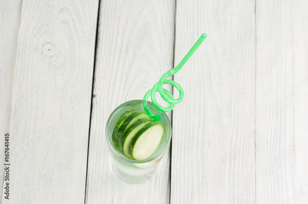 Many slices of green fresh cucumber in glass with water and green spiral plastic tubule on old white wooden planks Many slices of green fresh cucumber in glass with water and green spiral plastic tubule on old white wooden planks