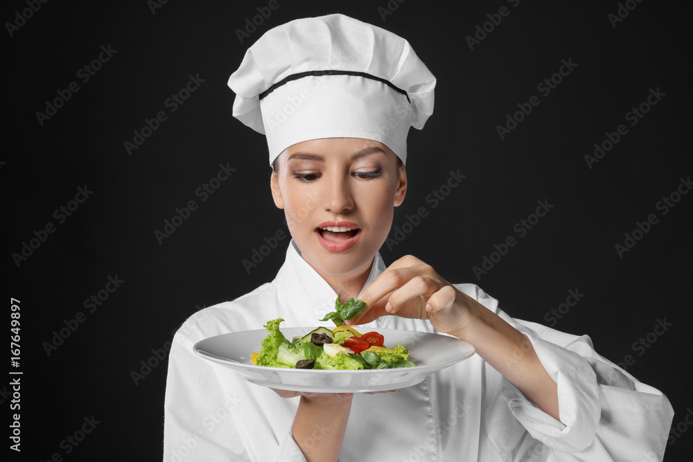 Young female chef holding plate with salad on dark background Young female chef holding plate with salad on dark background