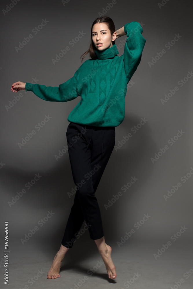 Attractive Caucasian happy sexy female model with brunette hair is moving in studio, wearing green sweater, isolated on dark background