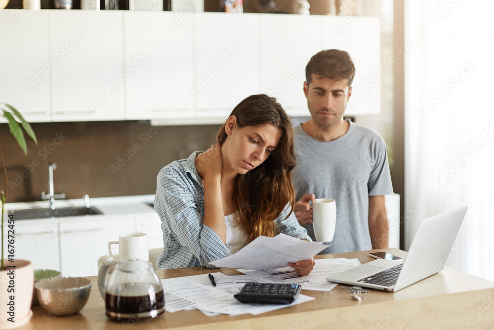 Φωτογραφία Worried adult Caucasian woman managing home accounts in kitchen, studying sheet of paper, her husband holding cup of coffee and looking over her shoulder