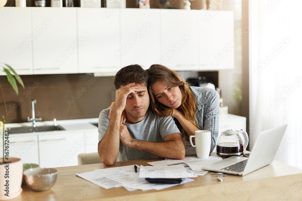 Stressed wife and husband with many debts doing paperwork together, reviewing their expenses, planning domestic budget and calculating bills in kitchen, surrounded with papers, calculator and laptop Stressed wife and husband with many debts doing paperwork together, reviewing their expenses, planning domestic budget and calculating bills in kitchen, surrounded with papers, calculator and laptop