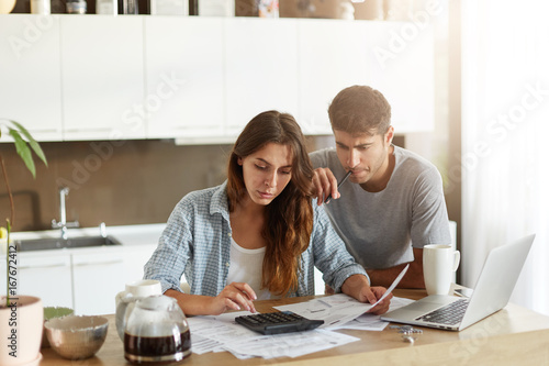Busy attractive young woman calculating something on calculator, holding documents in hands while her husband leaning at her shoulder, keeping pen in mouth being preoccupied with financial report