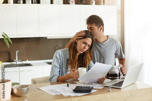 Frustrated woman studying sheet of paper in kitchen, drinking coffee and trying to find way to pay off all family debts; her husband kissing her on head, saying: Everything is going to be alright photo