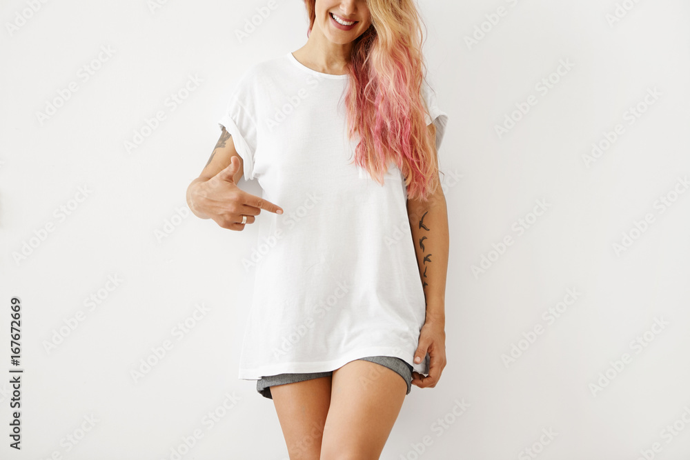 Φωτογραφία Slim woman with pink hair tips, wearing white loose T-shirt and jean shorts, pointing at copy space for your text message or promotional content, isolated over white studio background