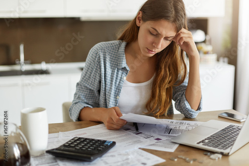Indoor shot of casually dressed young woman holding papers in her hands, calculating family budget, trying to save some money to buy new bicycle to her son, having stressed and concentrated look photo