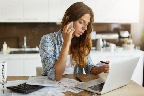 Beautiful young Caucasian woman calling bank using cell phone concerning information on credit card that she is holding. Serious female connecting to mobile banking service using electronic device