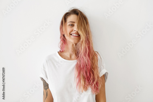 Horizontal portrait of pleasant-looking Caucasian female with long hair, pink on tips, having tattooes on arms, wearing white casual T-shirt, covering her face with hair, looking happily in camera photo