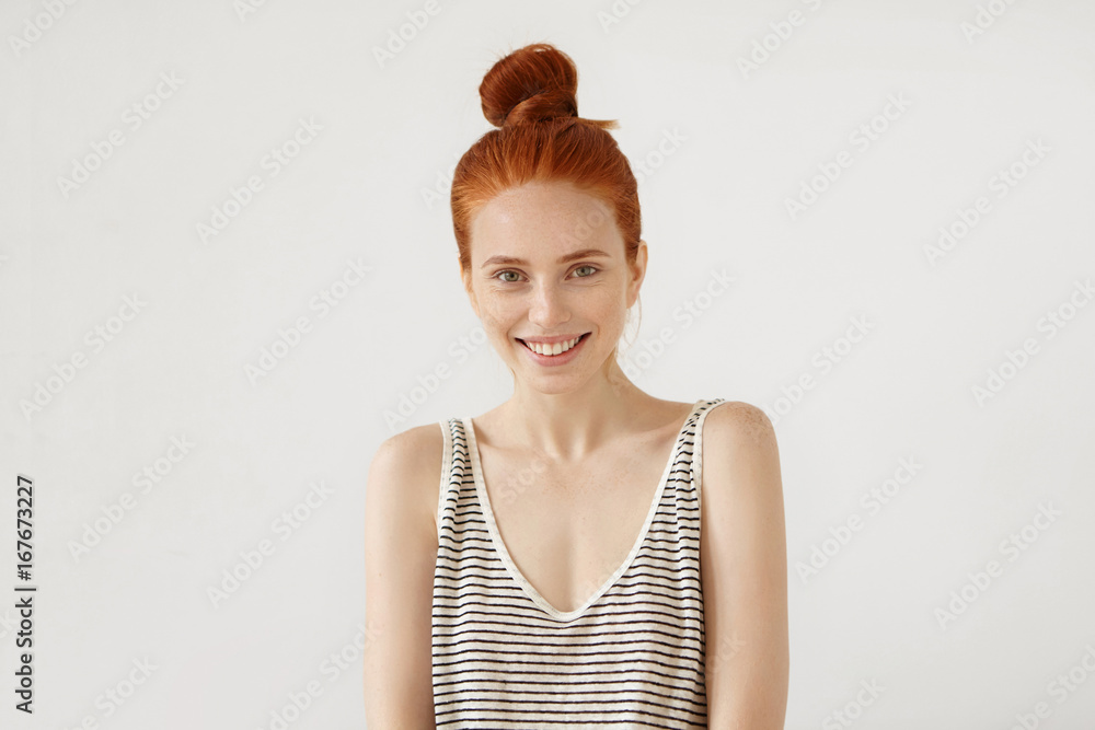 Φωτογραφία Happy freckled woman with red hair knot, wearing loose striped shirt, feeling relaxation while resting at home, having good mood, smiling pleasantly, showing her perfect white teeth