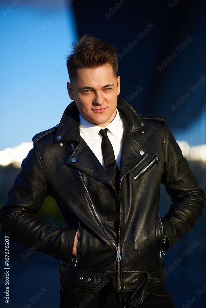 Urban style and street fashion concept. Outdoor picture of trendy-looking  smooth-shaven young Caucasian man wearing stylish leather jacket over white  shirt with black tie, posing with hands in pockets Stock Photo