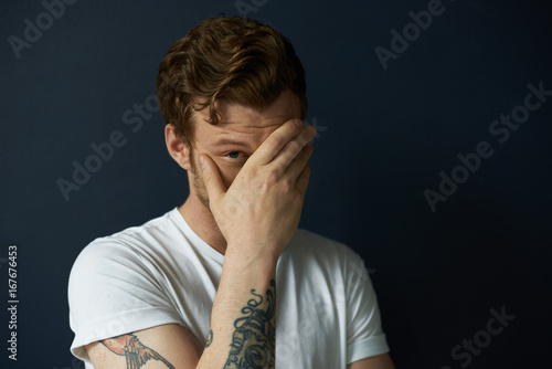 Masculinity concept. Headshot of handsome tattooed young European male fashion model dressed in white casual t-shirt looking out of his palm, flirting, having playful facial expression. Body language
