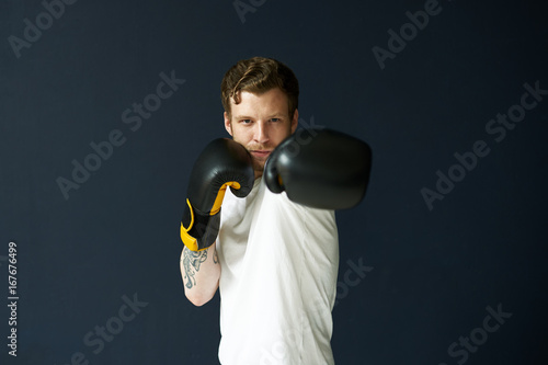 Indoor portrait of serious young unshaven European fighter in black boxing gloves working out in gym before competition, looking at camerawith confident expression on his face, ready for fight photo