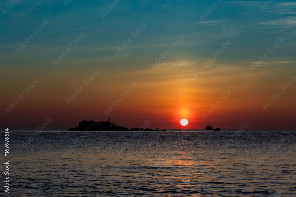 A fiery orange sunrise sky looking out over the south China sea in Nha Trang Bay Vietnam. With fishing boats and a small island in silhouette.