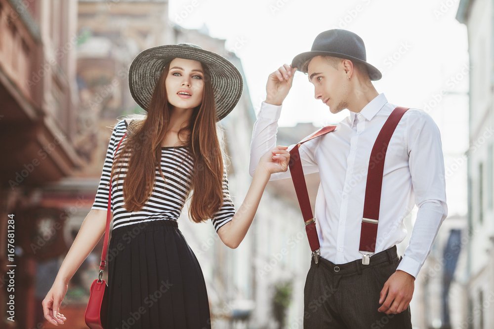 Outdoor portrait of young beautiful fashionable couple. Man and woman posing in street. Girl holding his suspenders. Models wearing stylish clothes and accessories. Sunny day light. Fashion concept
