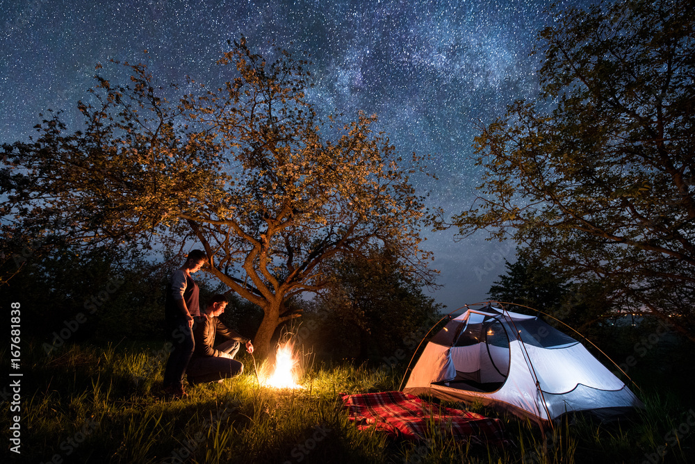 Romantic couple tourists standing at a campfire near tent under trees and beautiful night sky full of stars and milky way. Night camping - obrazy, fototapety, plakaty 