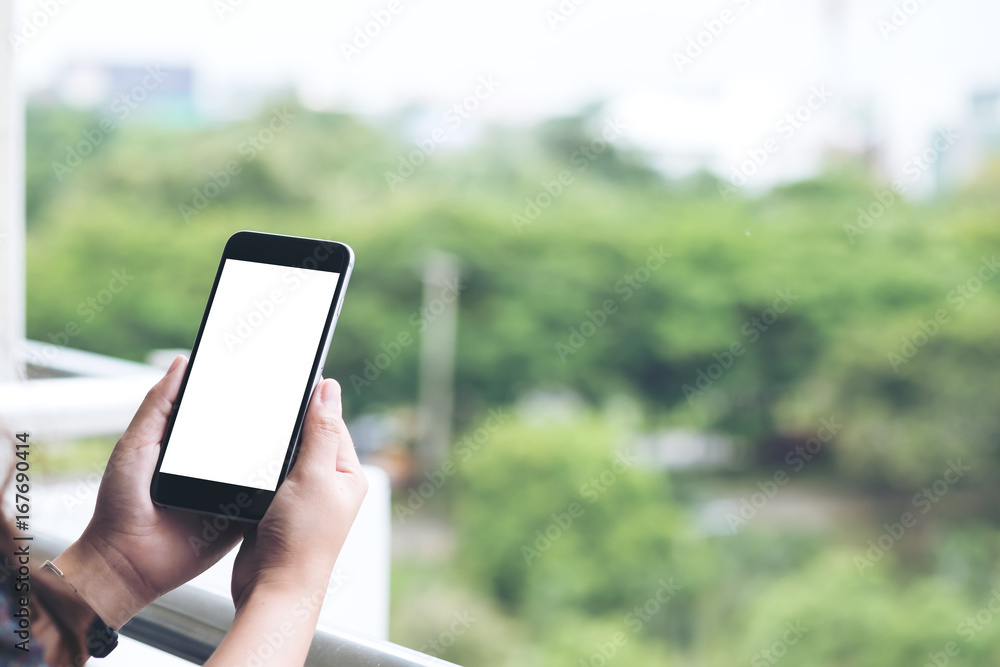Mockup image of a woman's hand holding and using black smart phone with blank white screen while standing at the balcony of a high building with metal rail and blur green nature background