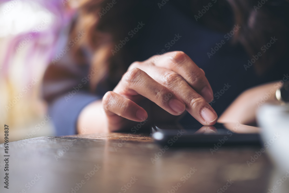A business woman's hand touching and sliding finger on a black smart phone on wooden table in modern cafe