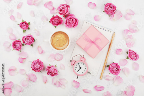 Morning coffee cup, gift box, alarm clock and pink rose flowers on white table top view in flat lay style. Greeting card for Mother or Woman day.