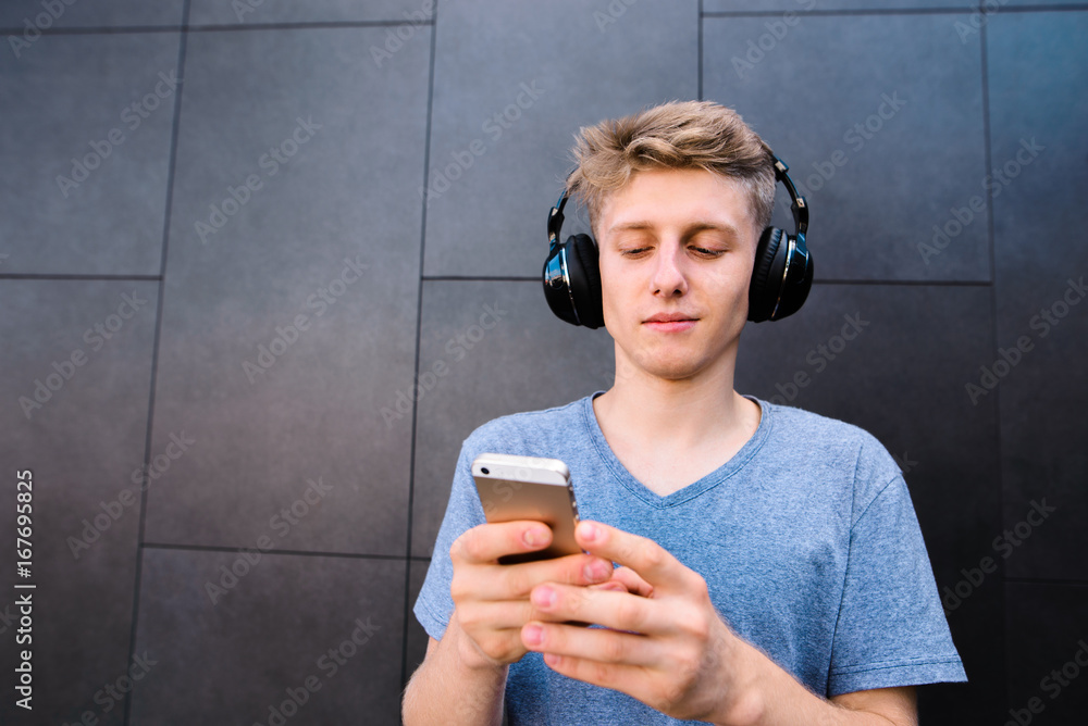 Fototapeta premium Portrait of a smiling young man listening to music in wireless headphones and using a phone on the background of a gray wall.
