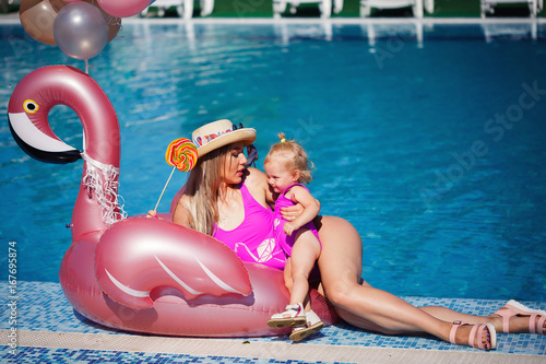 Mom and daughter in bright pink bathing suits rest on a pool with inflatable flamingos © endrews21
