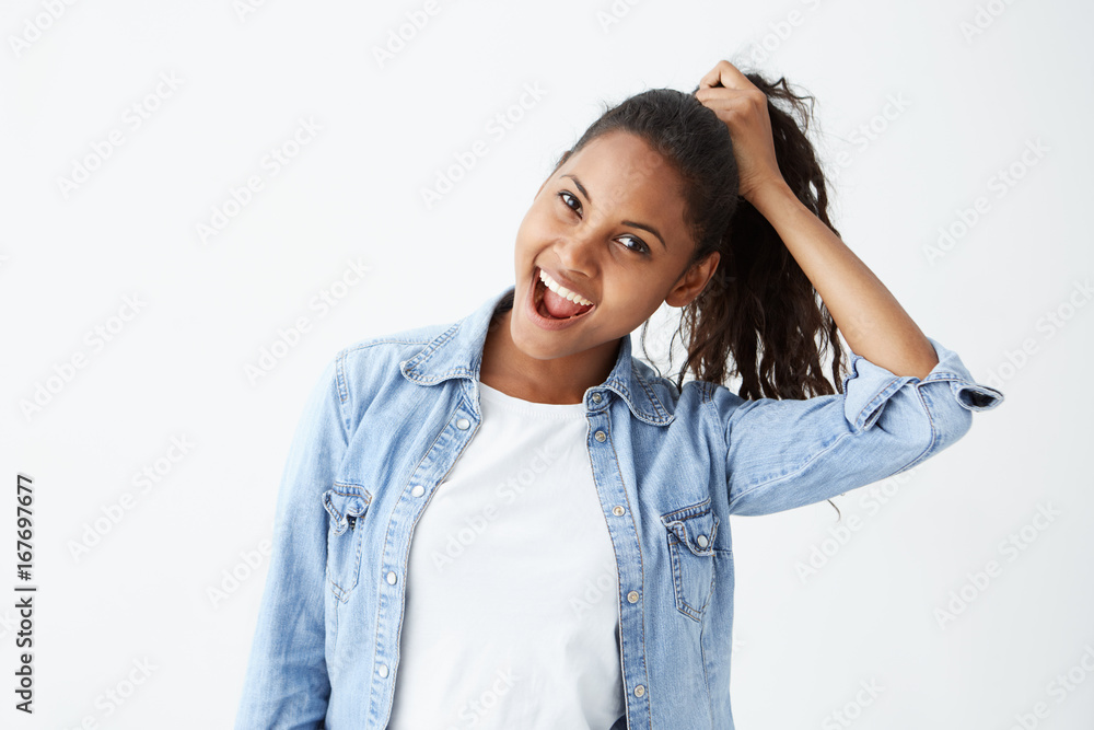 Human face expressions and emotions. Portrait of young African-American brunette woman in light-blue denim shirt having fun, holding her hair with hand, with mouth wide open. Cheerful and joyful Human face expressions and emotions. Portrait of young African-American brunette woman in light-blue denim shirt having fun, holding her hair with hand, with mouth wide open. Cheerful and joyful