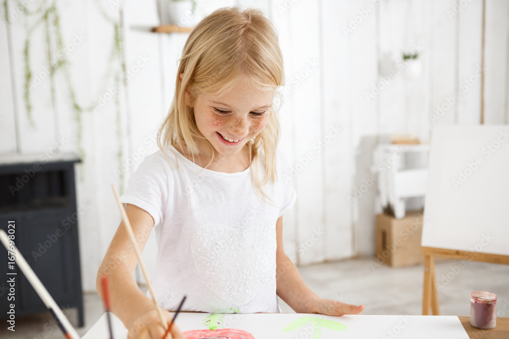 Smiling and cheerful, full of joy child with blonde hair and freckles holding brush in her hand and aspiringly painting picture at the art room. Shot of happy blonde little girl using watercolors