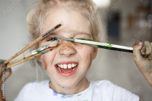 Blonde little girl in playful mood with paint on her freckled face and blue eyes covering her face with brushes and looking through them at you like hiding. Playing, smiling child showing her teeth