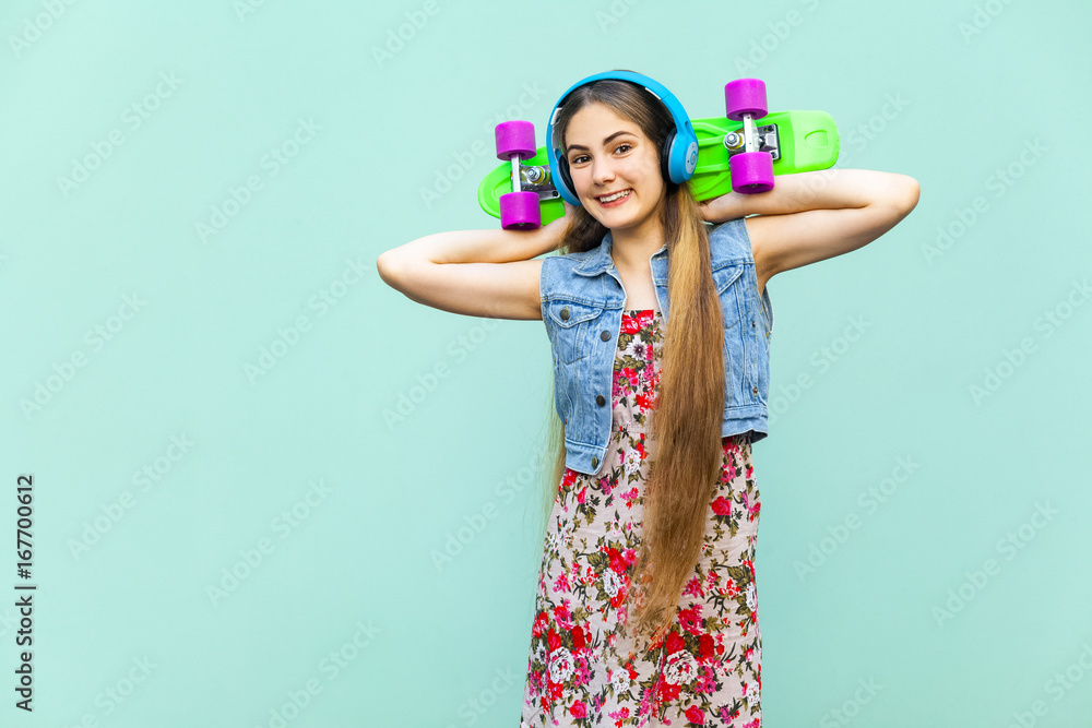 The young happy beautiful long haired blonde girl in dress and headphones, having fun with green plastic penny skateboard in front of the light blue wall. Isolated studio shot The young happy beautiful long haired blonde girl in dress and headphones, having fun with green plastic penny skateboard in front of the light blue wall. Isolated studio shot