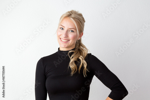 Junge Frau mit jugendlichem Aussehen im schwarzen engen Pullover vor einer weißen Wand mit schönen blonden langen gesunden Haaren photo