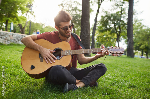Good-looking stylish bearded man casually dressed having pleased expression being glad to spend time outdoors playing guitar and singing songs. Hipster guy sitting cross-legged in park holding guitar.